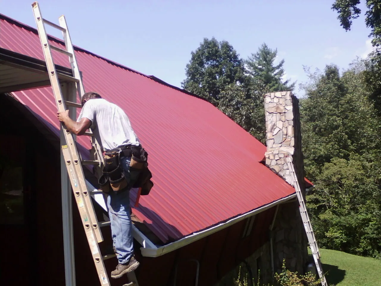 Roofer on a ladder beside a red metal roof and stone chimney — Soffit & Fascia Repair by Roofing Brothers in Friendship